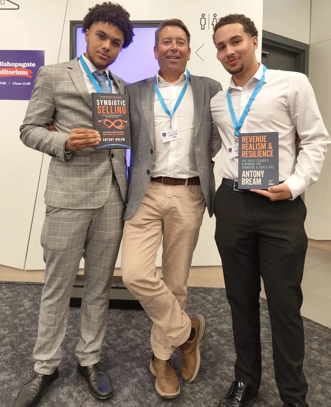 Two young founders with their mentor at a business event, holding copies of Symbiotic Selling and Revenue, Realism & Resilience
